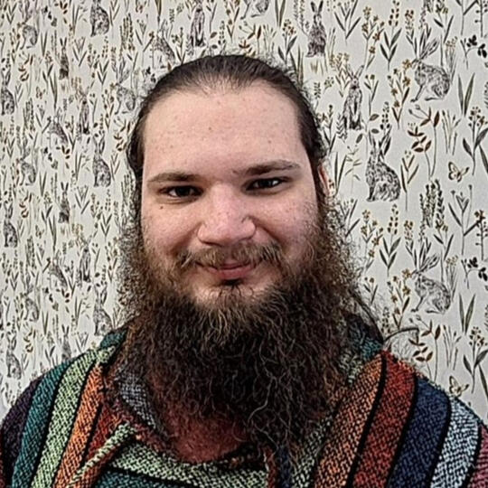 A photograph of Kieran, a white non-binary person with long, dark brown hair and beard and blue eyes. They are wearing a many coloured striped baja and behind them is a pale backdrop with many illistrations of hares amongst various grasses.